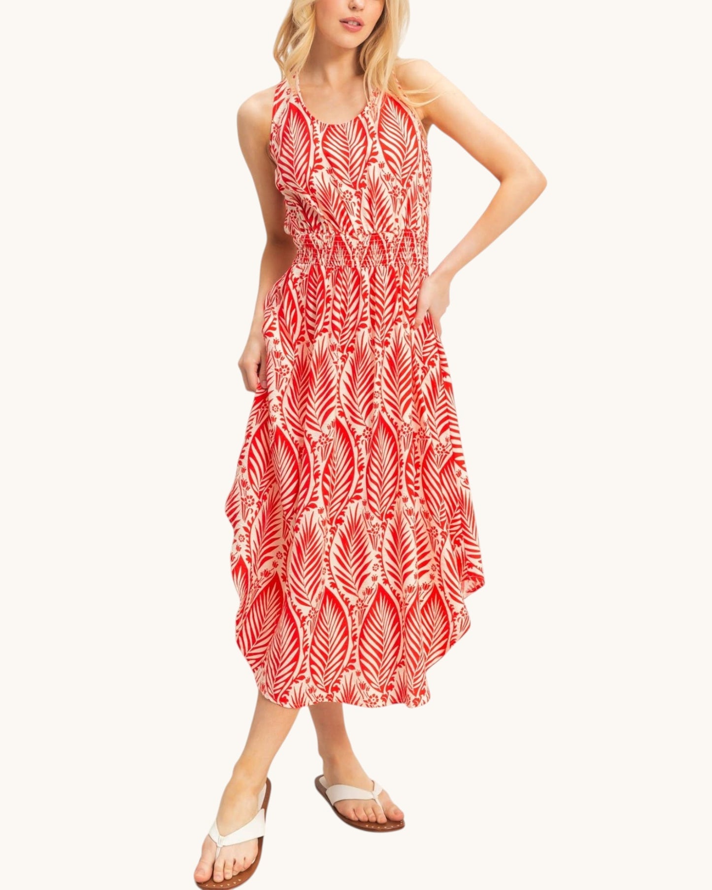 A woman modeling a sleeveless midi dress with a red and white leaf pattern, paired with white slide sandals, against a plain off-white background.