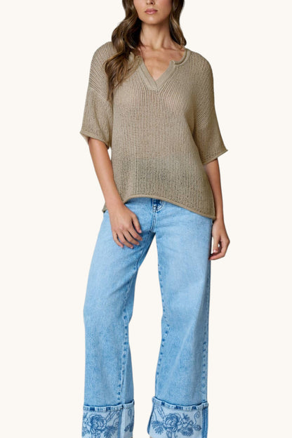 A woman wearing a loose, short-sleeve knit top in a cool taupe color paired with light blue wide-leg jeans featuring paisley-patterned cuffs, set against a solid off-white background.