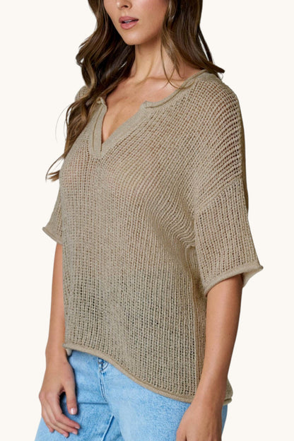 A woman wearing a loose, short-sleeve knit top in a light taupe-beige color with a split neckline, paired with light blue jeans against a plain off-white background.