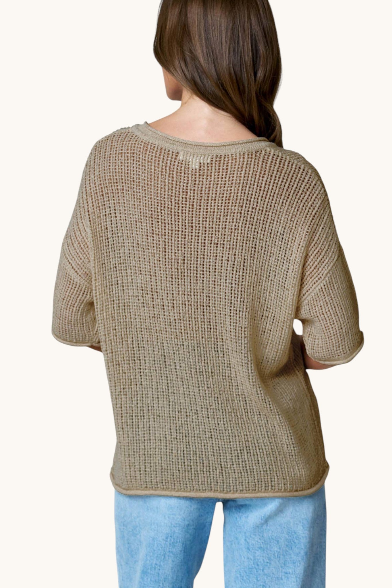 A woman viewed from the back wearing a loose, textured knit short-sleeve top in a cool taupe-beige color, paired with light blue jeans, against a plain off-white background.
