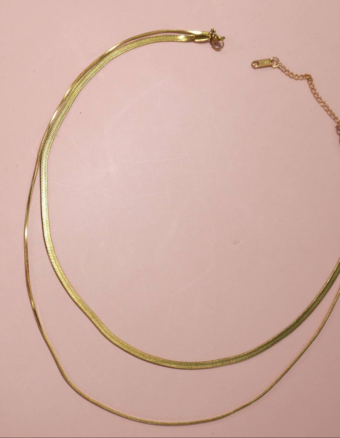 A double-strand gold snake chain necklace with a small clasp and extender chain, displayed on a solid salmon pink background.