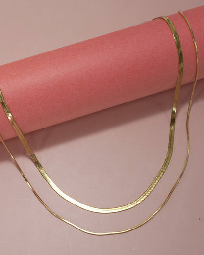 A thin gold chain necklace draped over a pink cylindrical display prop against a pink background.