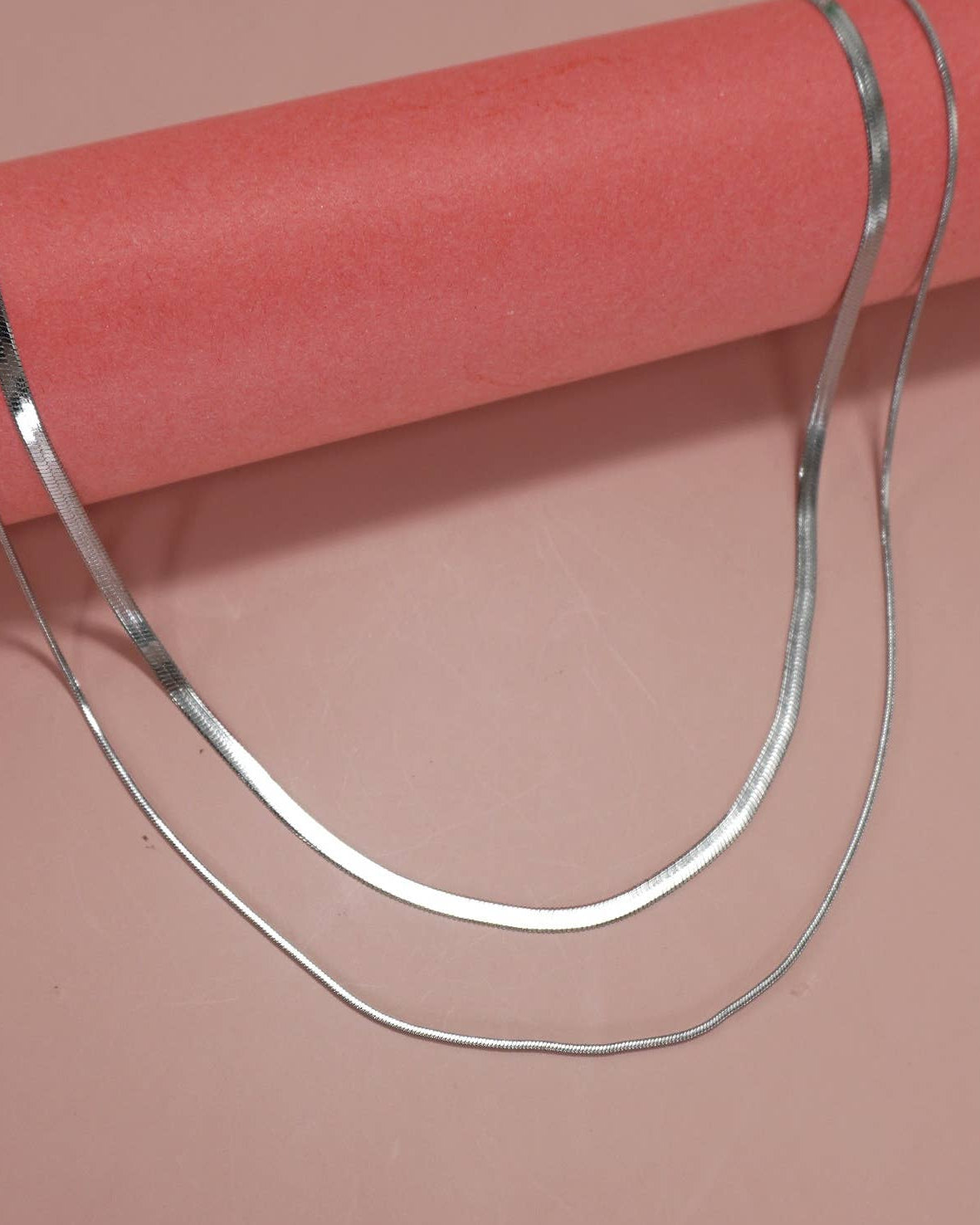 A double layer necklace in silver featuring a herringbone and snake chain draped over a pink cylindrical display stand against a solid salmon pink background.