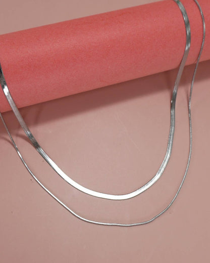 A double layer necklace in silver featuring a herringbone and snake chain draped over a pink cylindrical display stand against a solid salmon pink background.
