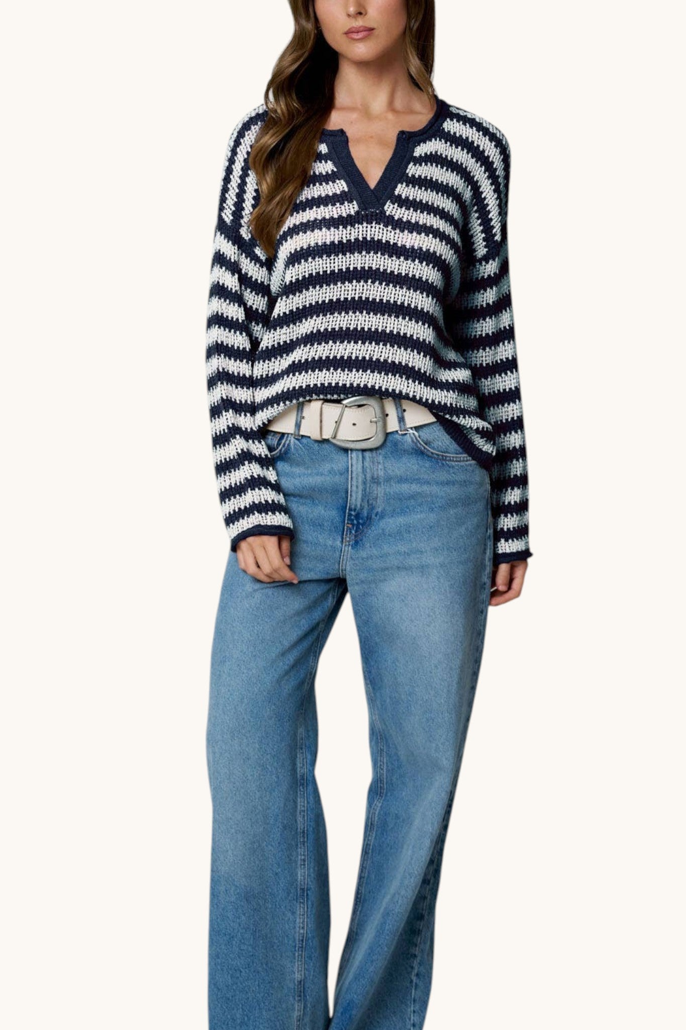A woman wearing a navy and white horizontally striped knit sweater with a split V-neck, styled with light-wash blue jeans and a silver belt, against a plain off-white background.