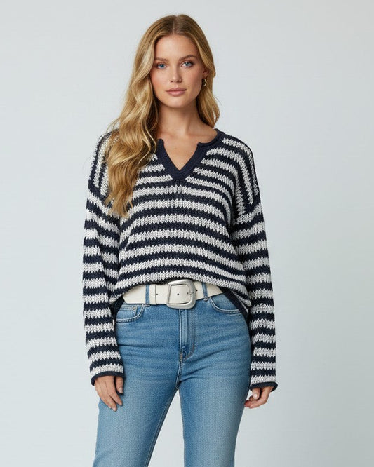 A woman modeling a navy and white horizontally striped knit sweater with a V-neck, paired with light blue jeans, a white belt, and white sneakers, against a light gray studio background.