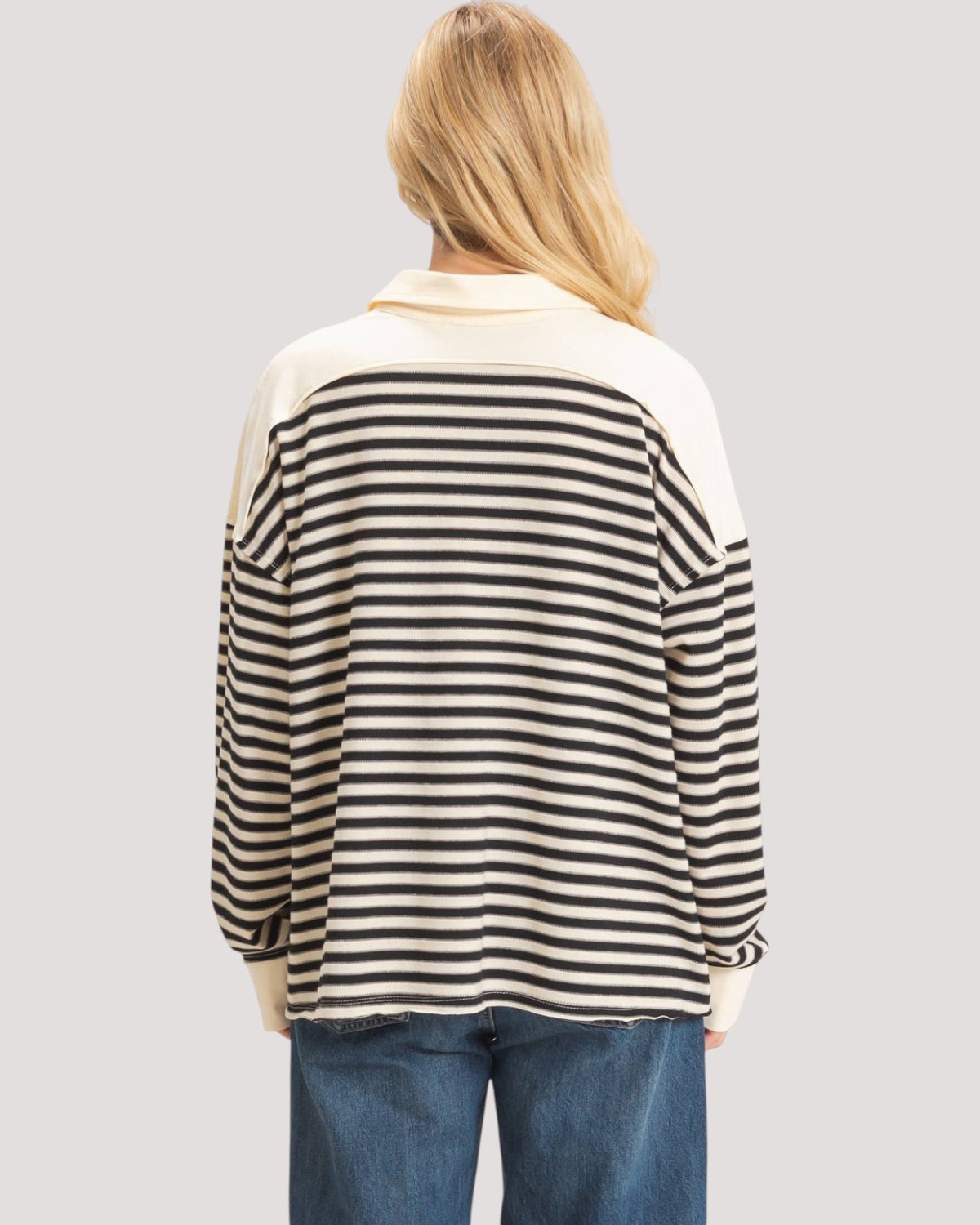A woman viewed from behind wearing a long-sleeve collared top with black and off-white horizontal stripes across the body and solid off-white sleeves and collar, paired with blue jeans against a light gray studio background.