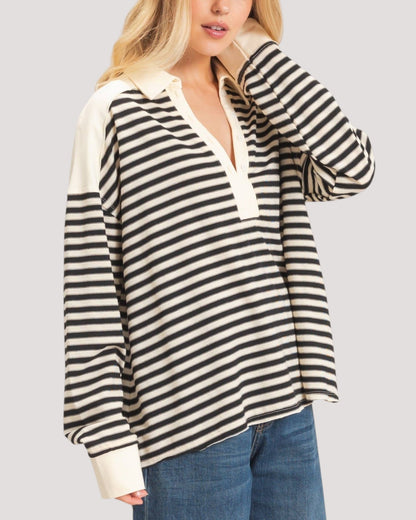 A woman wearing a black and off-white horizontally striped long-sleeve polo shirt with solid off-white shoulder panels and collar, paired with blue jeans, against a solid light gray background.