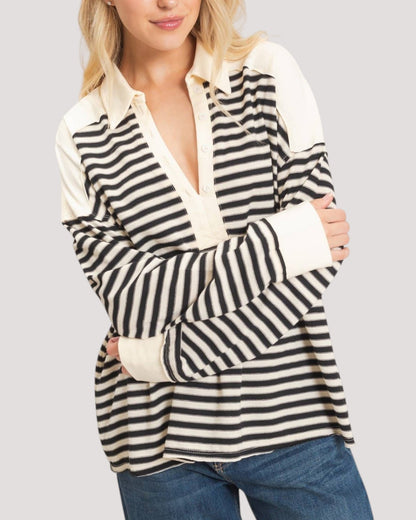 A woman wearing a long-sleeve rugby-style shirt with black and off-white horizontal stripes and solid off-white shoulder panels and collar, paired with blue jeans against a light gray studio background.