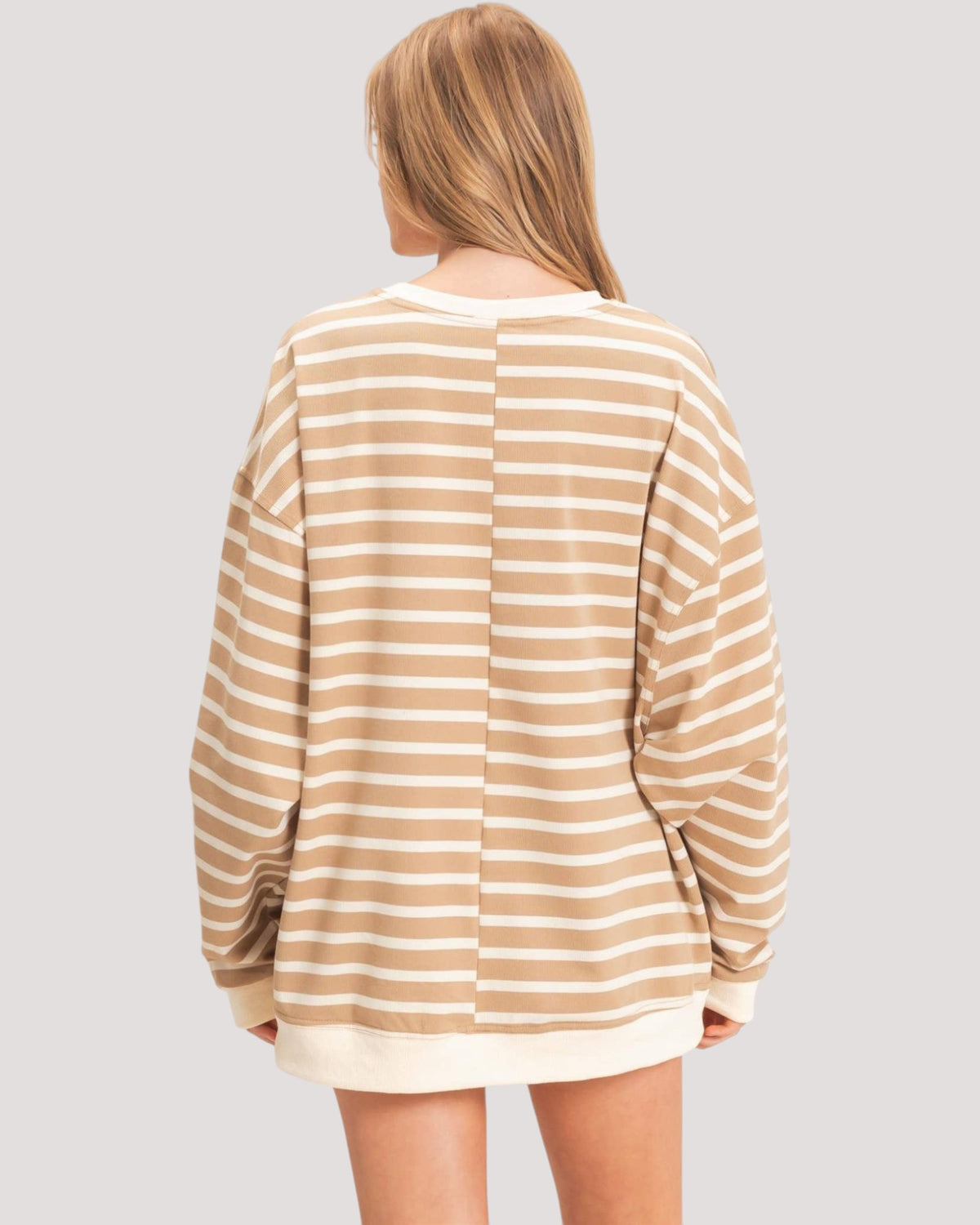 A woman viewed from the back wearing a loose, long-sleeve striped sweatshirt with horizontal beige and off-white stripes, set against a solid light gray background.