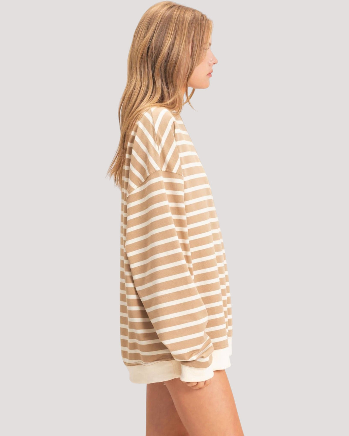 A woman shown in side profile wearing an oversized long-sleeve sweatshirt with horizontal beige and off-white stripes, photographed against a plain light gray studio background.
