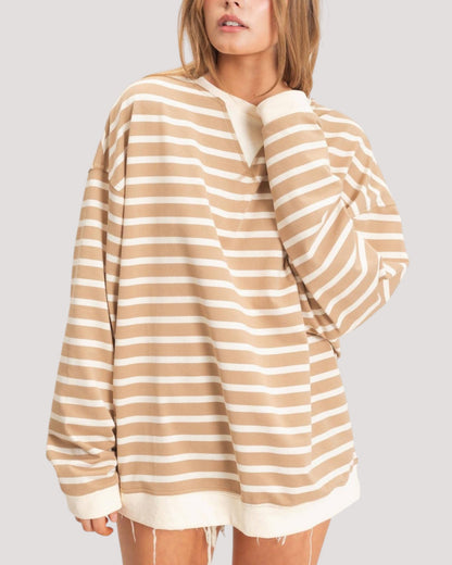 A woman wearing an oversized long-sleeve striped top with beige and off-white horizontal stripes, shown against a plain light gray studio background.