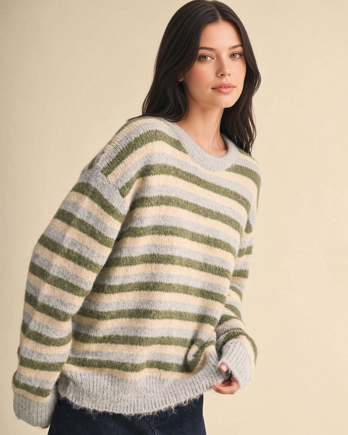 A woman wearing a fuzzy knit crewneck sweater with horizontal stripes in sage green, cream, and light gray-blue, posed against a solid beige studio background.