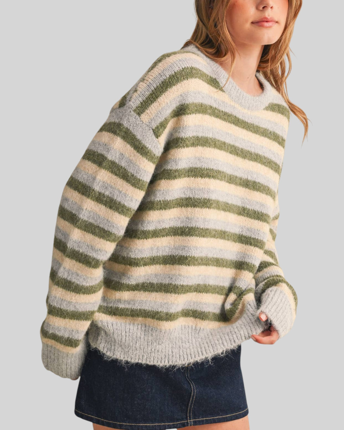 A woman wearing a fuzzy knit sweater with horizontal stripes in sage green blue-gray and cream, paired with a dark denim skirt, against a solid light gray background.