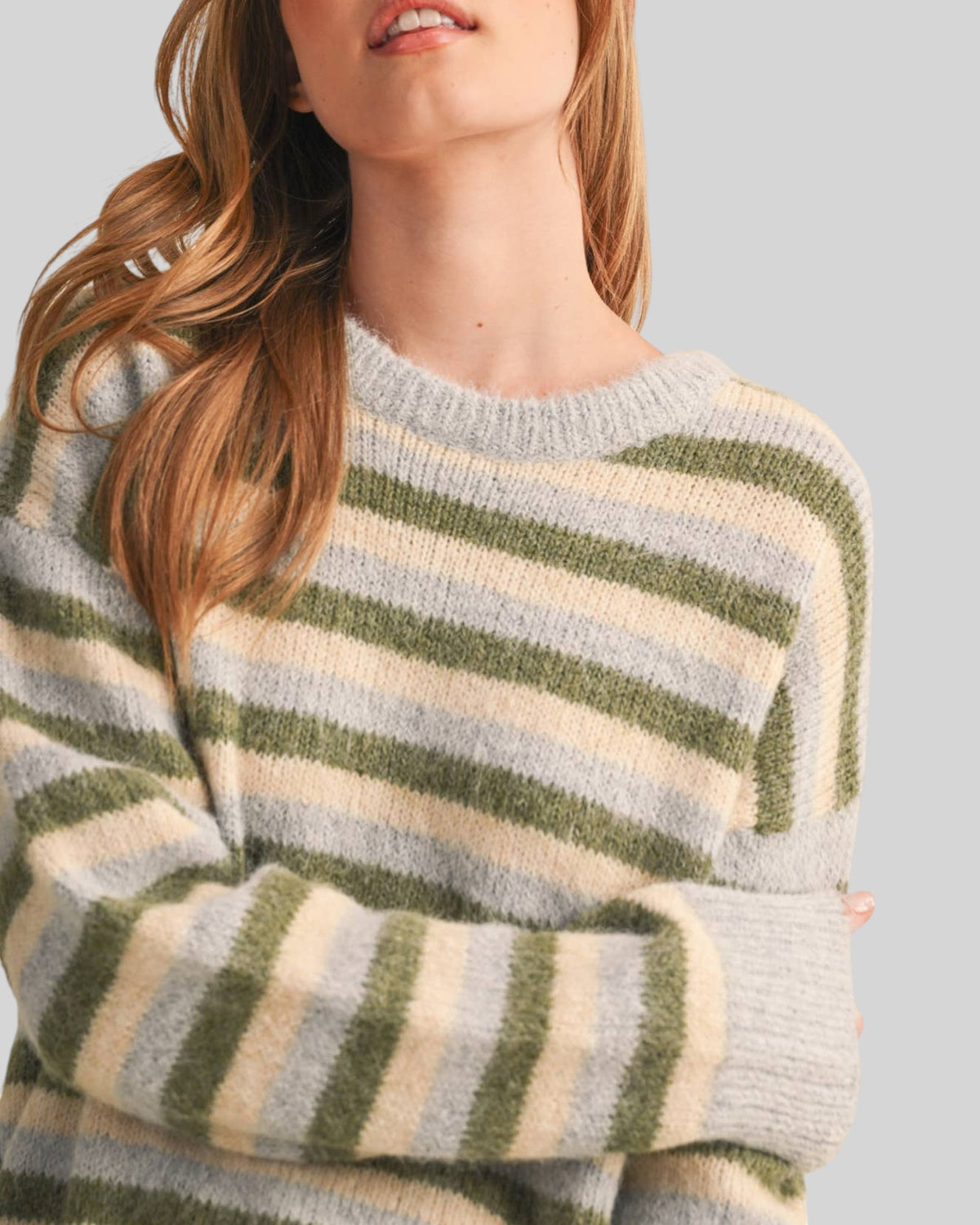 A close-up of a woman wearing a fuzzy knit sweater with horizontal stripes in sage green, cream, and light gray-blue against a solid light gray background.