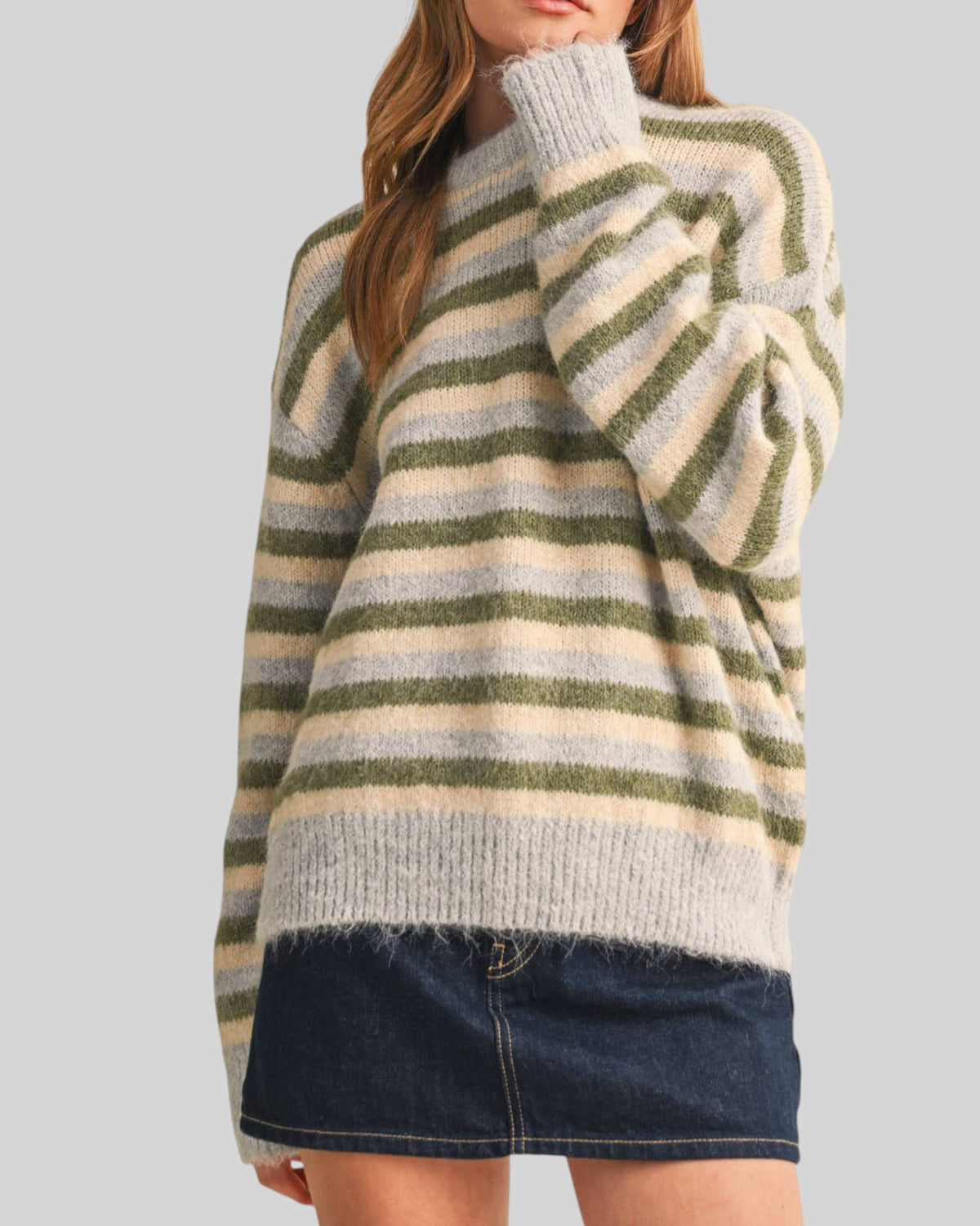 A woman wearing a fuzzy knit sweater with horizontal stripes in sage green, blue-gray and cream, paired with a dark denim mini skirt, against a solid light gray background.