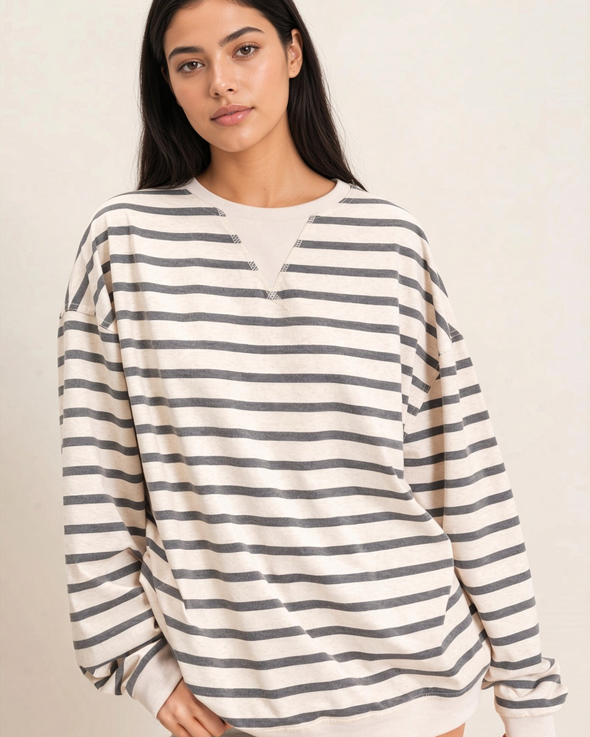 A woman wearing a long-sleeve crewneck sweatshirt with horizontal gray stripes on an off-white background, photographed against a plain light beige wall.