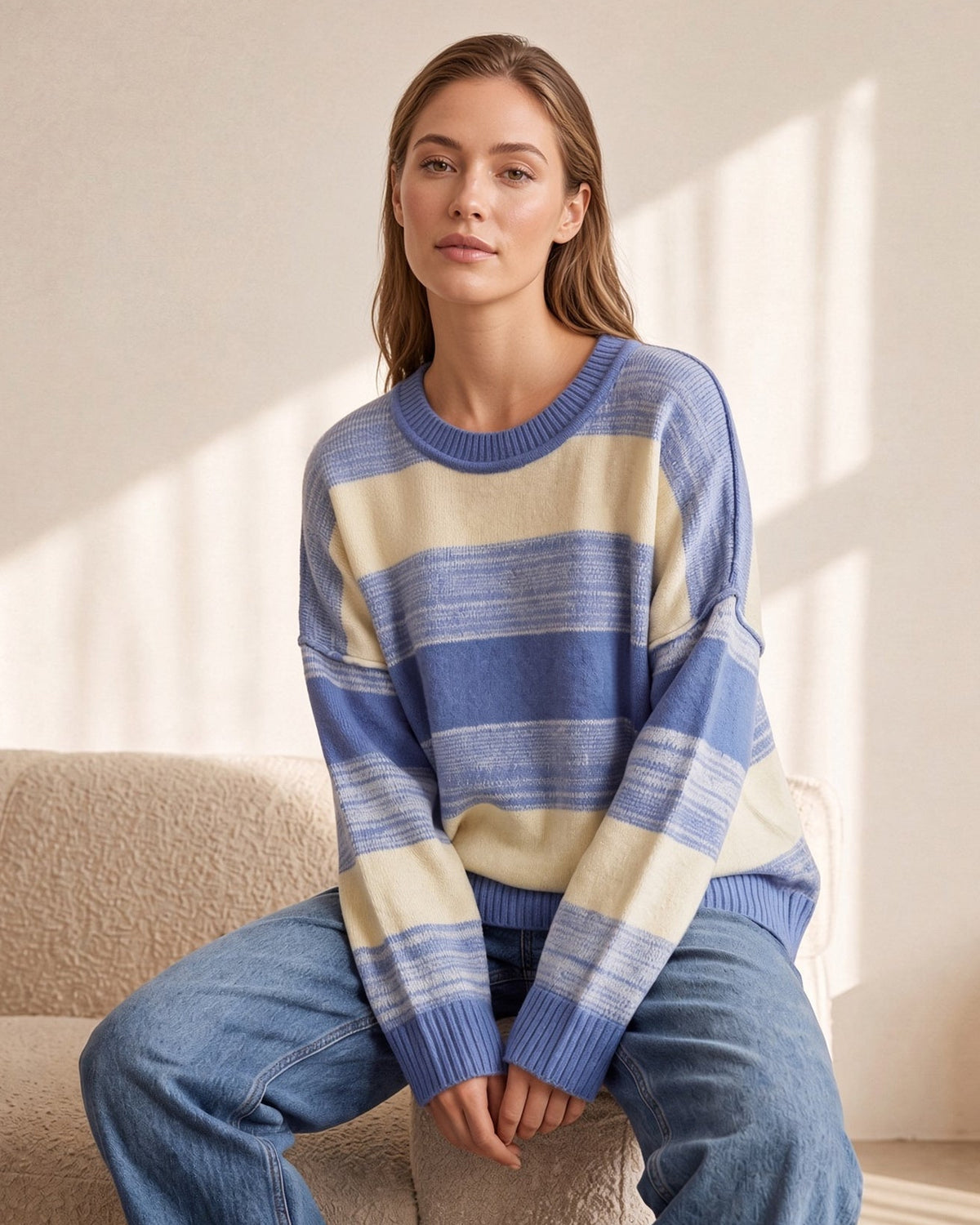 Seated model wearing blue and cream striped crew neck sweater with ribbed cuffs and jeans.