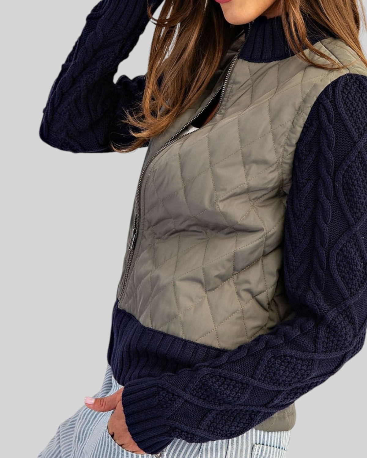 Side view of a woman wearing a Navy and Olive mixed-media jacket with quilted body, navy cable knit sleeves, and ribbed trim with a zipper front.