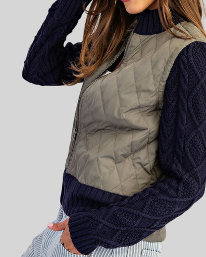 Side view of a woman wearing a Navy and Olive mixed-media jacket with quilted body, navy cable knit sleeves, and ribbed trim with a zipper front.