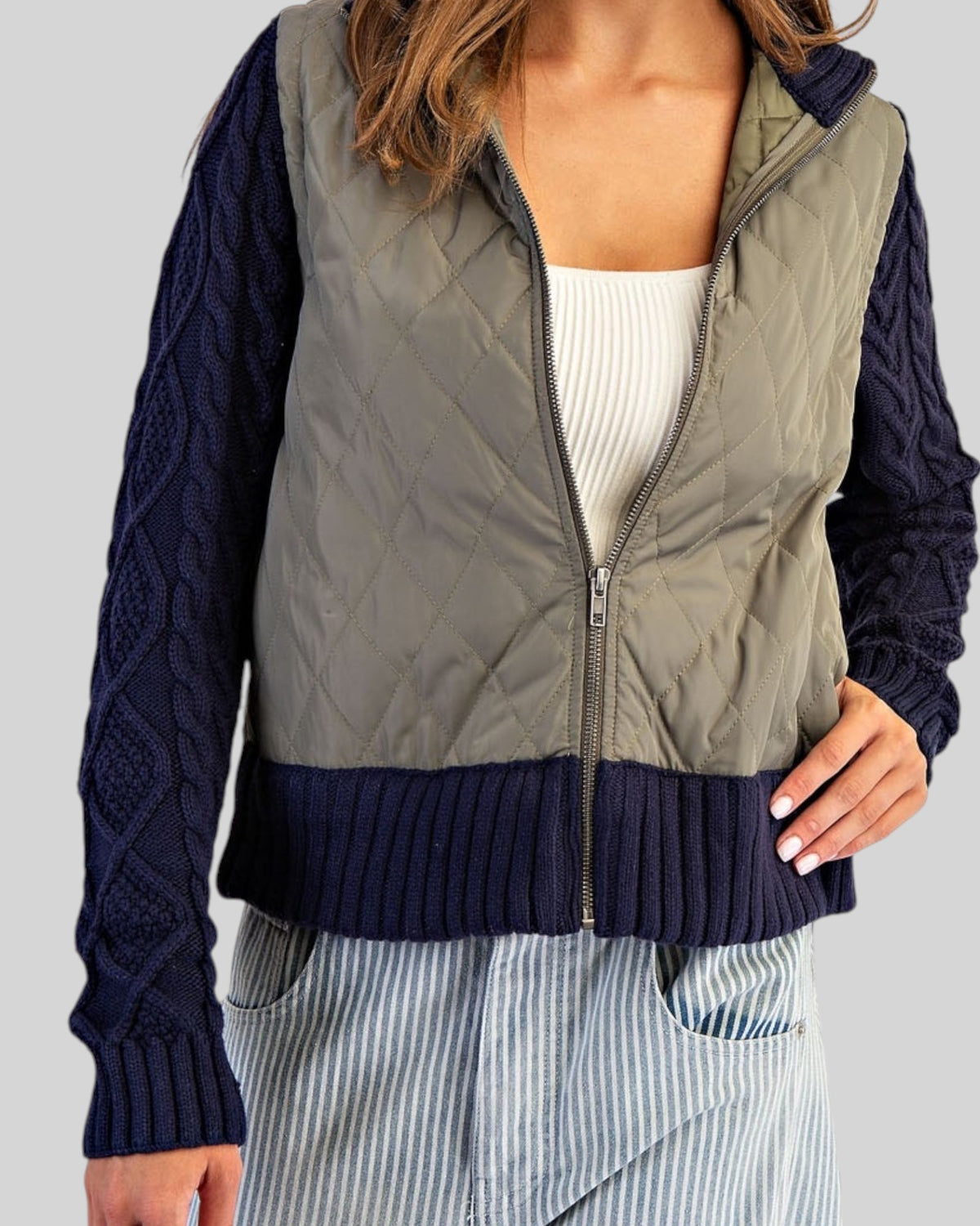 Navy and olive mixed-media jacket with quilted body, navy cable knit sleeves, and ribbed trim with a zipper front worn with a white tee shirt.
