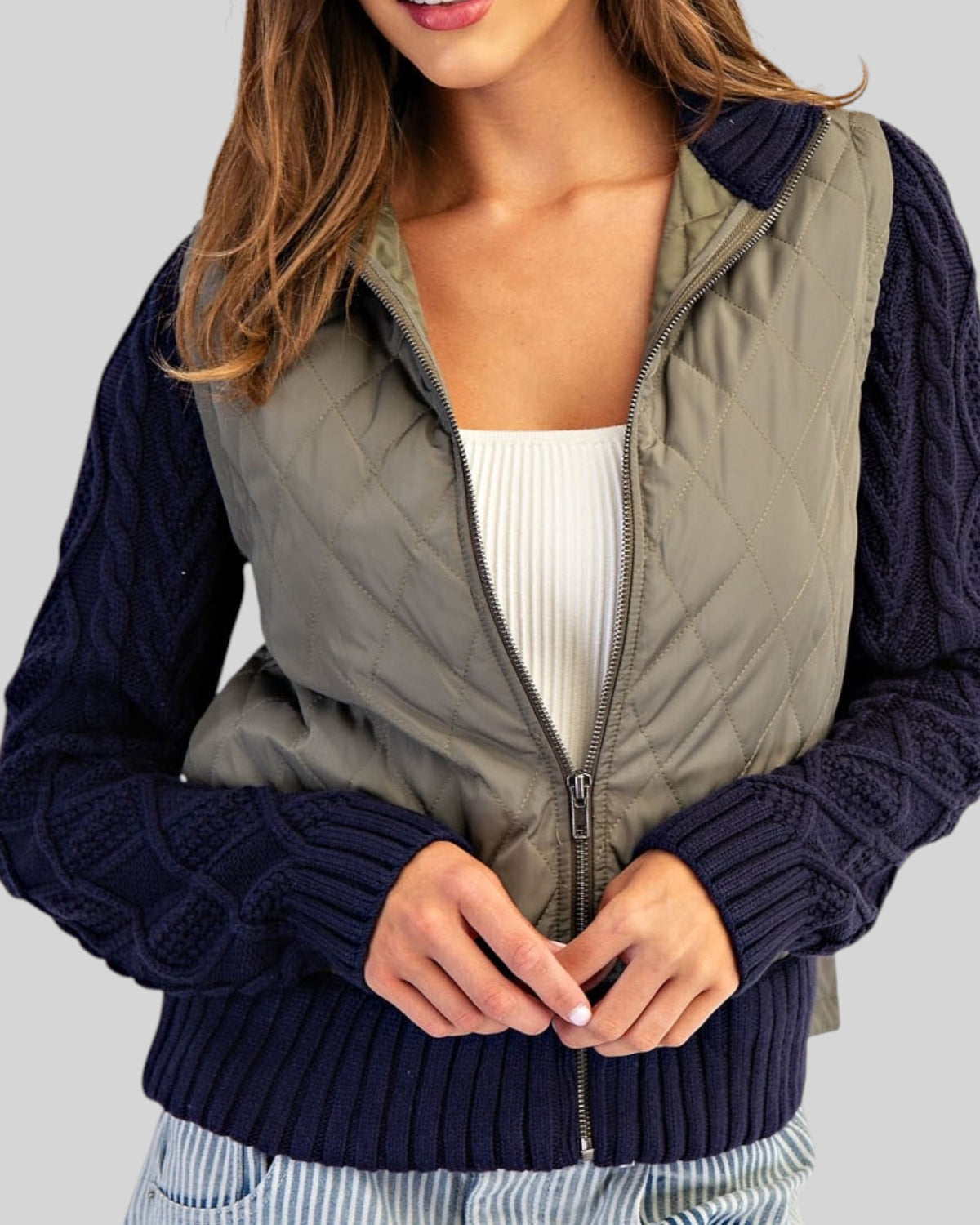 Navy and olive mixed-media jacket with quilted body, navy cable knit sleeves, and ribbed trim with a zipper front worn with a white tee shirt.