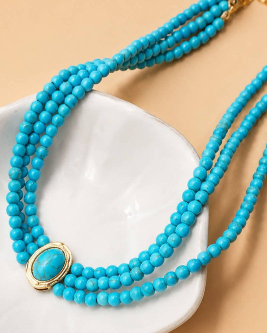 A multi-strand turquoise beaded necklace with a gold oval bezel setting, displayed on a white ceramic dish against a warm beige background.