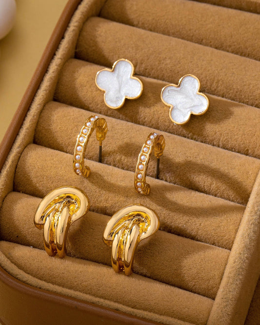 Gold and white enamel clover earrings, pearl huggie hoops and gold knot earring set in a jewelry box on a beige background.