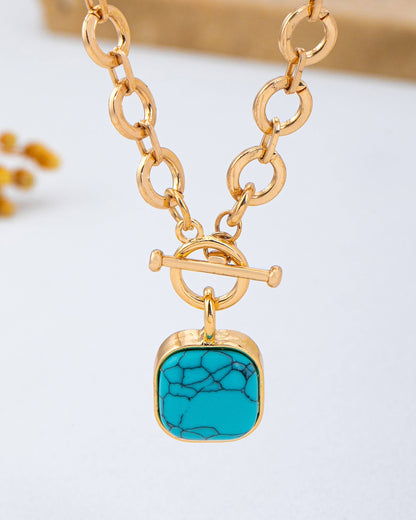 Close up of a gold chain toggle necklace with a turquoise pendant on a light background.